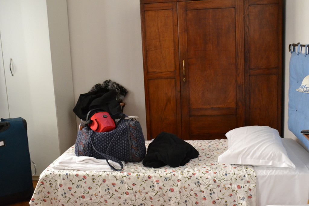 A neatly made bed with a floral bedspread in a room, with a large wooden wardrobe in the background and a bag with belongings placed on the bed.