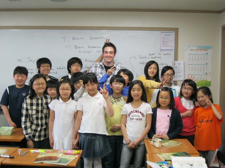 A classroom of smiling students posing with their English teacher in front of a whiteboard filled with notes, showcasing a diverse group of children in a cheerful learning environment.