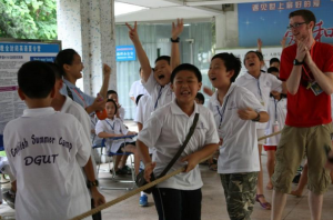 china-summercamp | Abroad101 Blog A group of campers in white “English Summer Camp DGUT” shirts cheer and laugh in an indoor hallway in China; one boy runs toward the camera holding a broom handle while a staff member in a red shirt claps nearby.