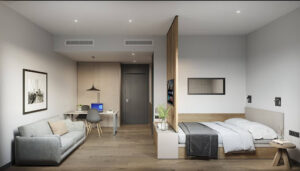 Blau 1 | Abroad101 Blog Modern studio apartment interior at BLAU Student Housing & Language Academy in Barcelona, featuring a combined living and sleeping area. The space is well-lit, with wooden flooring, a gray sofa, a study desk with a blue chair, a bed with a gray comforter, and minimalist decor. A large, sliding wooden partition partially separates the bed from the rest of the room.
