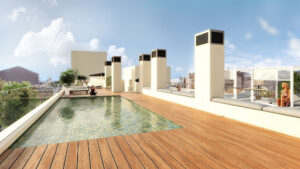 Blau 2 | Abroad101 Blog Rooftop pool area at BLAU Student Housing & Language Academy with wooden decking. The pool has clear blue water and is flanked by white walls and modern, square-shaped pillars. Lounge chairs are placed beside the pool for relaxation. The deck offers a view of surrounding buildings under a bright blue sky. To the right, there's a transparent safety barrier through which a person is visible, enjoying the sunshine.