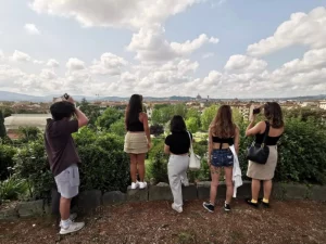 A group of five young adults attending the SRISA study abroad program on a hilltop, observing and taking photos of the panoramic view of Florence, with historic cityscapes under a cloudy sky.