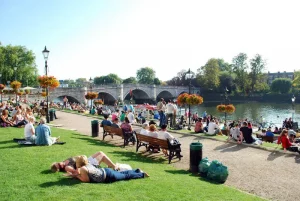 St Mary's University Twickenham, Greater London - Summer School program photo | Abroad101 Blog A lively summer scene by the river at Twickenham, with people relaxing and socializing on the grass and benches, near St Mary's University, with hanging flower baskets and the arching Richmond Bridge in the background.