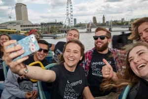Students from the University of Westminster's study abroad program joyfully taking a selfie with the London Eye and the Thames River in the background. They are wearing University of Westminster t-shirts and smiling broadly, showcasing the lively and engaging atmosphere of their study abroad experience in London, UK. 