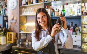 Free-bartending-schools-classes | Abroad101 Blog Smiling young woman practicing bartending skills while studying abroad, shaking a cocktail shaker behind a bar filled with various drinks and glasses.