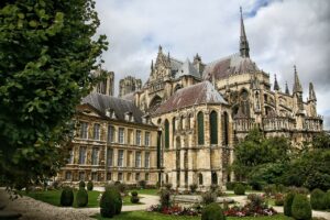 d | Abroad101 Blog Exterior view of Reims Cathedral in France, showcasing its intricate Gothic architecture with surrounding gardens and greenery under a cloudy sky.