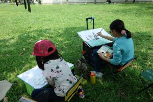 arthur-tseng-rWLyPPEo4M0-unsplash | Abroad101 Blog Two children sitting on small stools in a grassy park, focused on painting at portable easels during an outdoor art lesson.
