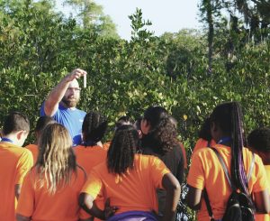 the-tampa-bay-estuary-program-IcUQNzaHrvg-unsplash | Abroad101 Blog Teacher leading a group of students in bright orange shirts on an outdoor lesson, holding up a test tube sample amid greenery to demonstrate hands-on learning in nature.