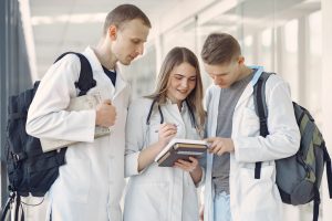 Three healthcare students in white lab coats and backpacks review notes together in a bright campus corridor—illustrating collaboration during a study‑abroad program.