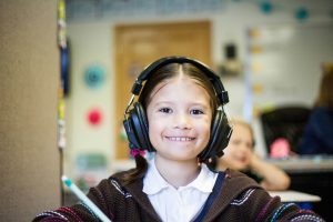Smiling elementary student wears oversized headphones in class, demonstrating audio learning tools that support focused, tech‑enhanced study.