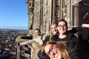APA burgundy visit | Abroad101 Blog Four APA study-abroad students smile on the balcony of a Gothic cathedral tower in Burgundy, France, overlooking the rooftops and countryside on a clear day.