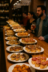 Perspective view of a long tapas counter inside a Basque pintxos bar; white plates stacked with bite-size baguette slices topped with various ingredients and toothpicks, a man holding a beer glass at right, blurred wine bottles and hanging glasses in the background.