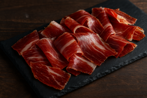 Close-up view of thin Jamón Ibérico de Bellota slices laid on a rectangular black slate serving board atop a wooden table; deep red meat with delicate white fat ribbons illustrates this premium Spanish cured ham.