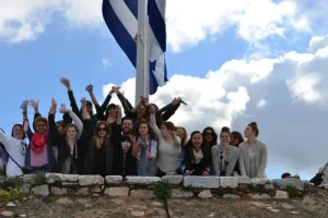 Program_1586_ProfilePhoto__64ef 1 | Abroad101 Blog ACT study-abroad students crowd a stone terrace, arms raised, beneath a tall Greek flagpole against a bright sky.