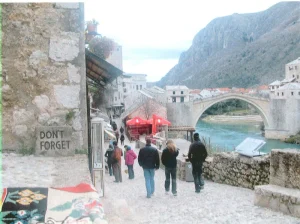 Program_488_Photo__a24a | Abroad101 Blog Visitors walking along a scenic cobblestone street with a bridge in the background, framed by mountains. A sign on the wall reads'DON'T FORGET,' reminding travelers of the area's history and significance.
