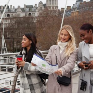 unnamed (6) | Abroad101 Blog Three young women who appear to be study abroad students walk together across a pedestrian bridge in a European-style city. The student in the center, wearing a light gray coat and scarf with a crossbody bag, holds an unfolded city map and smiles as she navigates. To her left, a student in a plaid coat carries a takeaway coffee and looks ahead, suggesting they are sightseeing or heading to a local destination. To the right, a student in a gray coat looks down at her phone while a compact camera hangs from her neck, capturing the feel of travel and cultural immersion. Historic buildings and winter trees fill the background, reinforcing the idea of exploring a new place, building confidence, and using everyday language skills to get around during a study abroad experience.