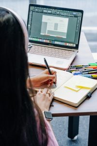 julio-lopez-1ZJRMbQrj64-unsplash | Abroad101 Blog Teacher (seen from behind) takes notes in a notebook beside an open laptop showing lesson materials, with pens and highlighters on the desk—representing ongoing professional development.