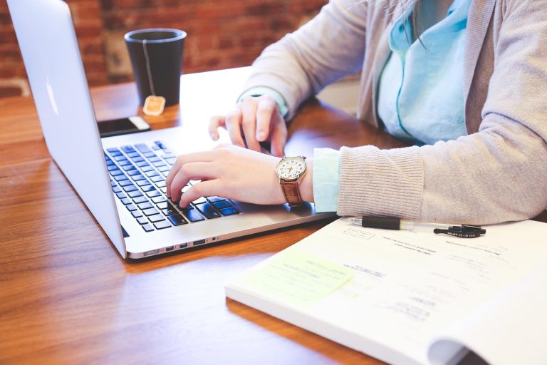Student typing on a laptop at a desk with notes and a coffee, planning coursework and adapting study habits while abroad.