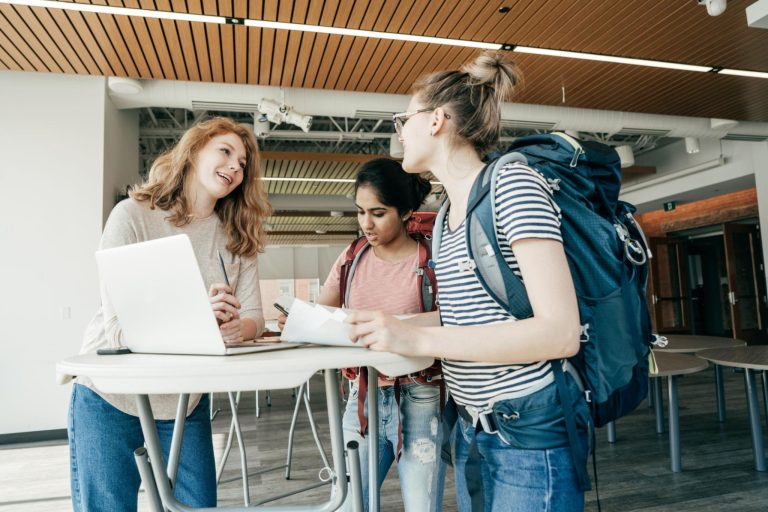 AmberStudent 1 | Abroad101 Blog Three students collaborate at a round table with a laptop and papers in a modern campus setting, backpacks nearby.