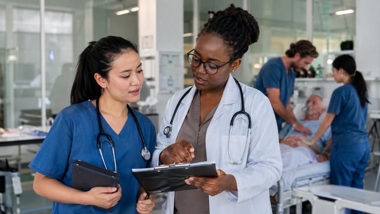 Medical trainee in scrubs discusses patient care with a doctor in a modern clinical training room.