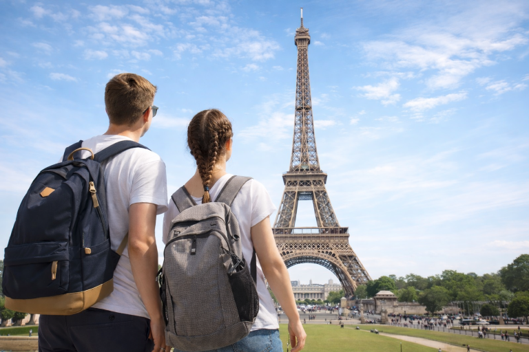 wo students with backpacks stand facing the Eiffel Tower in Paris on a bright day.