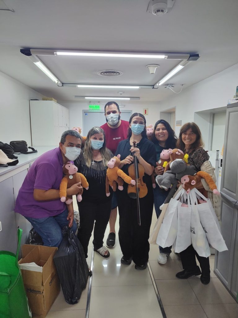 Group of people wearing masks in a bright hallway, holding stuffed monkeys and bags for a charity event; a violinist stands center.