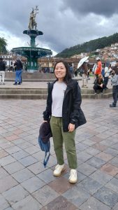 Studying Abroad with IPSL at Cusco, Peru | Abroad101 Blog Student standing in Cusco’s main plaza with a fountain and surrounding hills under cloudy skies.