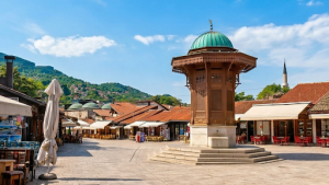 Hungary The Affordable Heart of Central Europe | Abroad101 Blog The ornate Sebilj wooden fountain stands in the centre of Baščaršija Square in Sarajevo, surrounded by outdoor cafés, traditional red-roofed buildings and green hills beneath a bright blue sky.