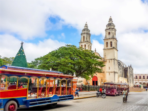Mexico Vibrant Culture in Our Backyard | Abroad101 Blog Colourful tourist trams travel through a historic Mexican plaza in front of a colonial cathedral with twin bell towers, while pedestrians and motorcyclists move through the square.