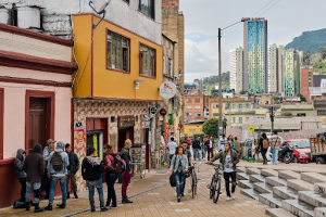 South Americas New Star is Colombia | Abroad101 Blog Pedestrians and cyclists gather along a colourful street in Bogotá, Colombia, with traditional neighbourhood buildings, modern high-rise towers and green mountains visible in the background.