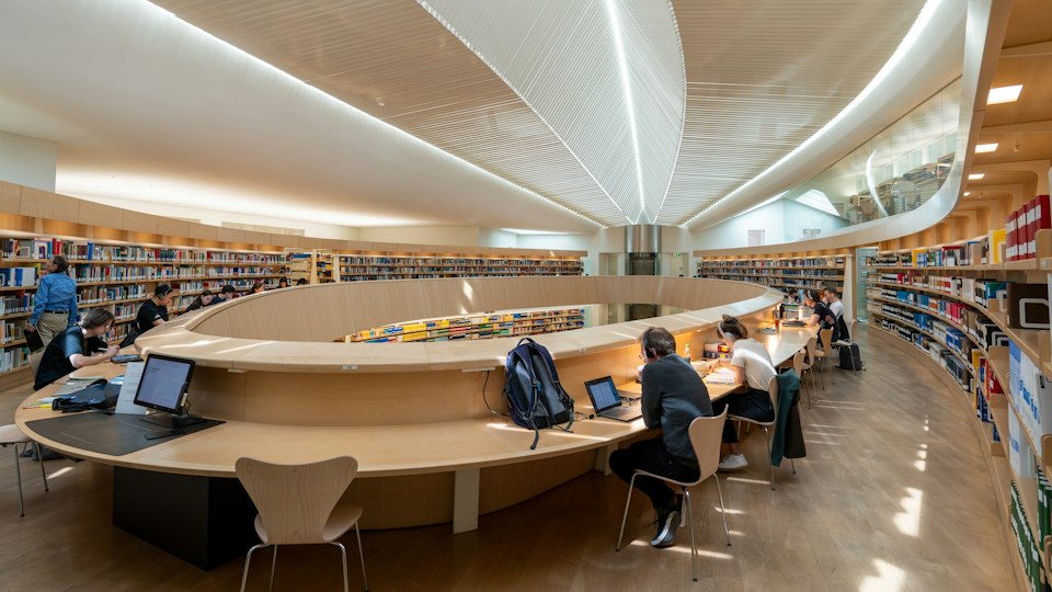 People studying at tables inside a large library lined with bookshelves.